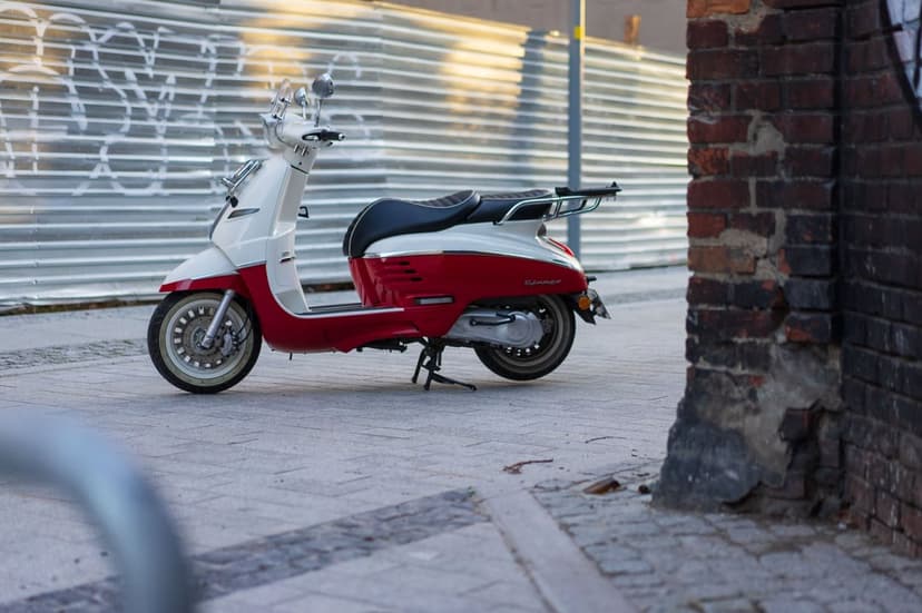 Red and white retro scooter parked on a stone sidewalk against a corrugated metal fence and brick wall.