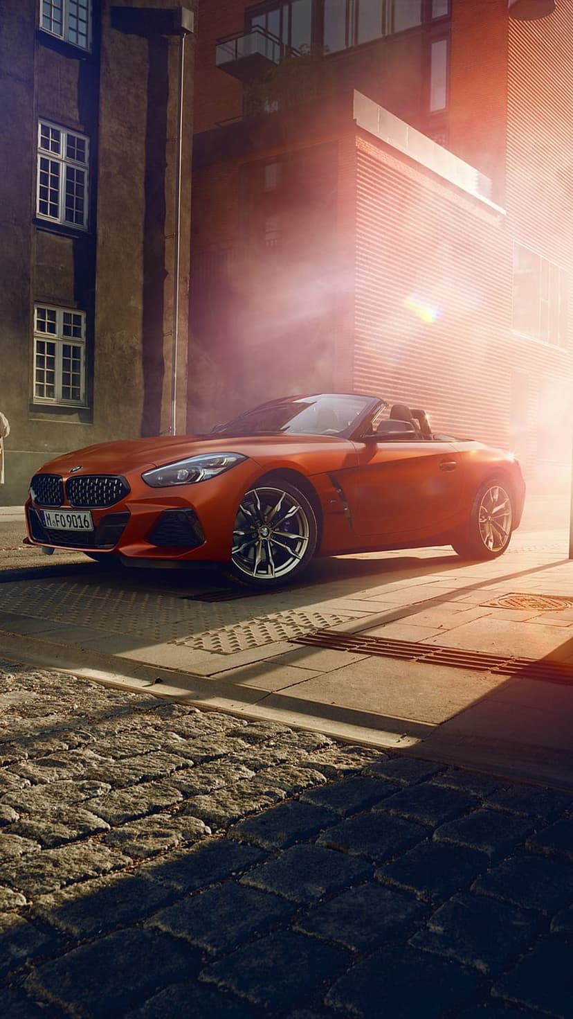 BMW Z4 Roadster in vibrant orange parked on a sunlit cobblestone street against a backdrop of modern buildings.