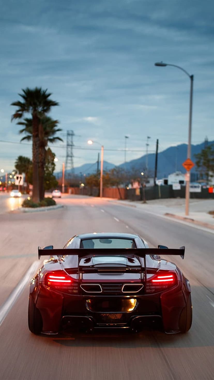 Red McLaren sports car with a large rear wing drives down a road lined with palm trees during a purple sunset.