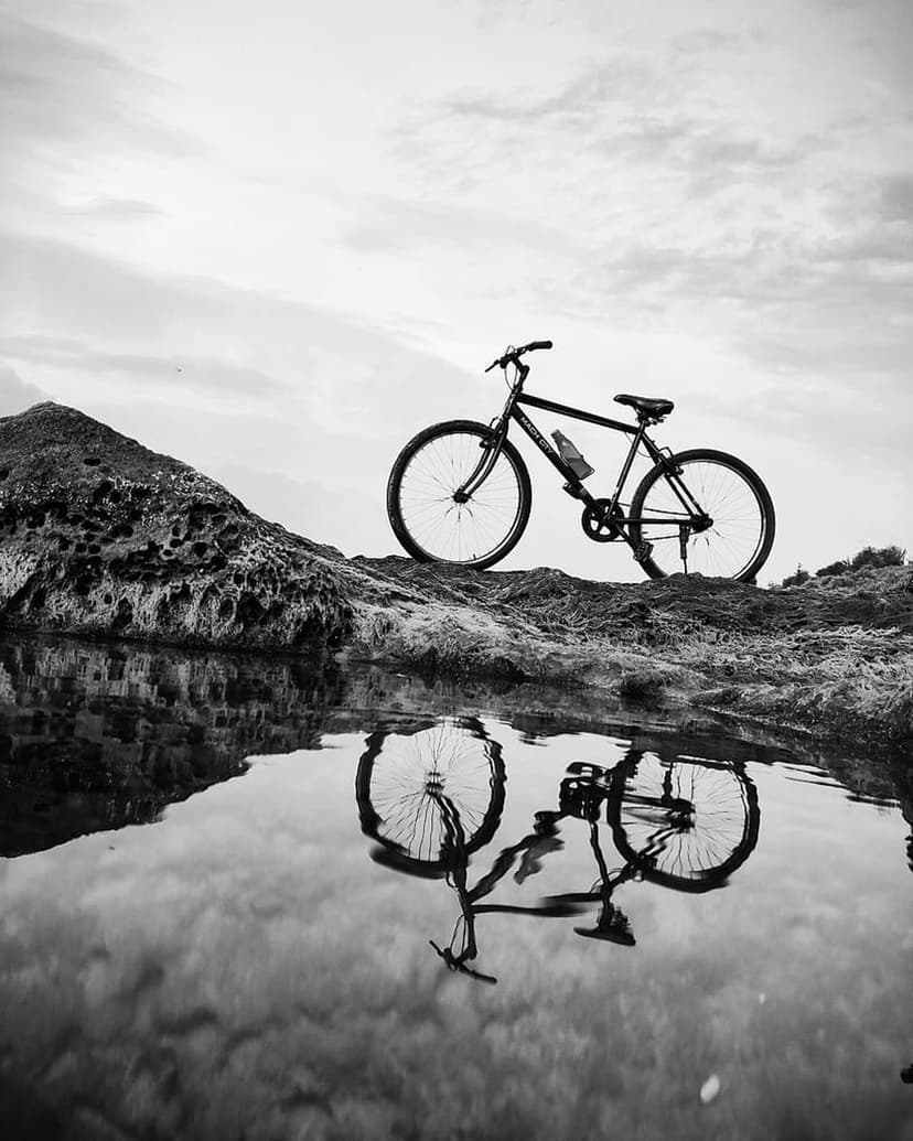 Vintage bicycle parked on a jagged rock formation reflecting in calm water under a dark, cloudy sky.