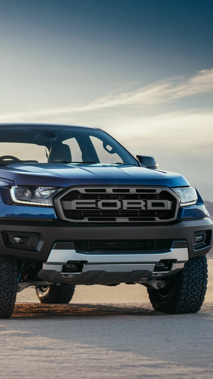 Front view of a rugged blue Ford Ranger Raptor pickup on desert sand dunes under a soft blue and orange dusk sky.
