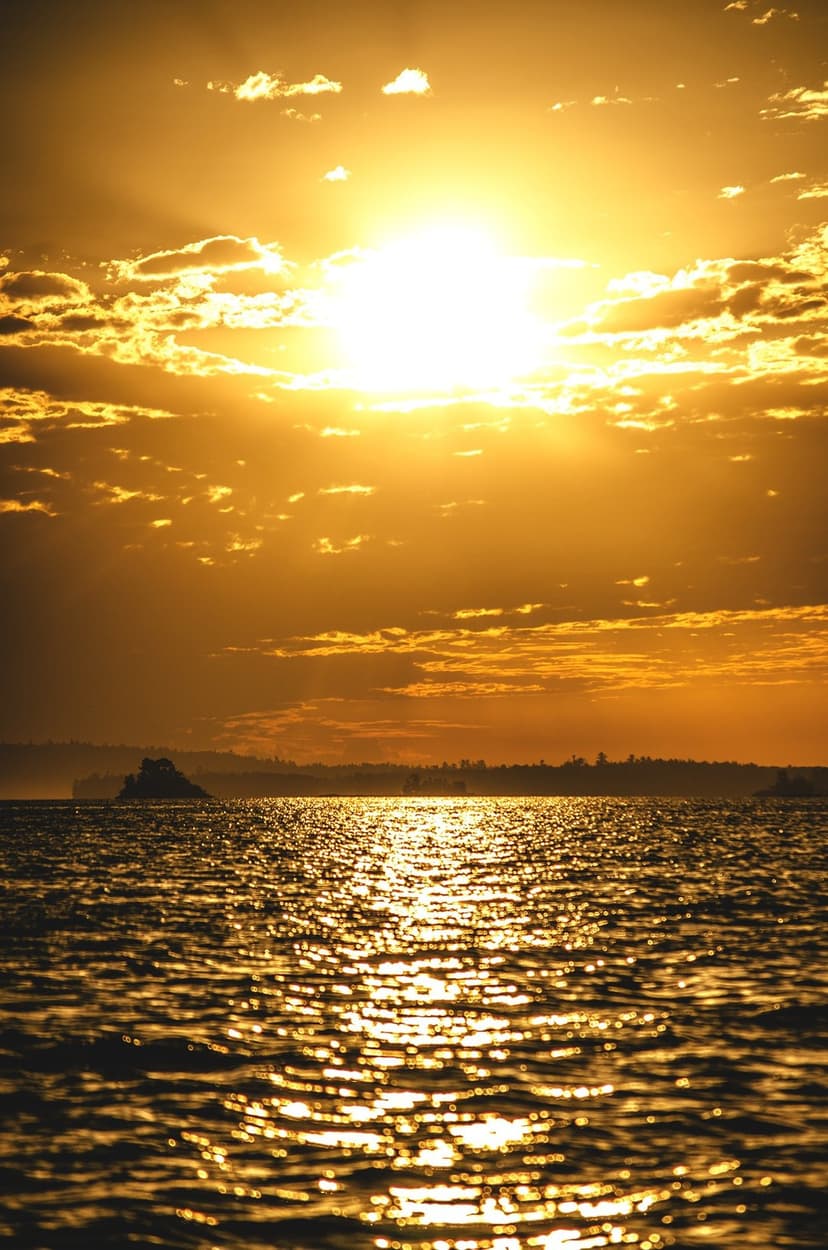 Golden sun setting over a calm sea with a dark island silhouette and textured orange clouds in the sky.