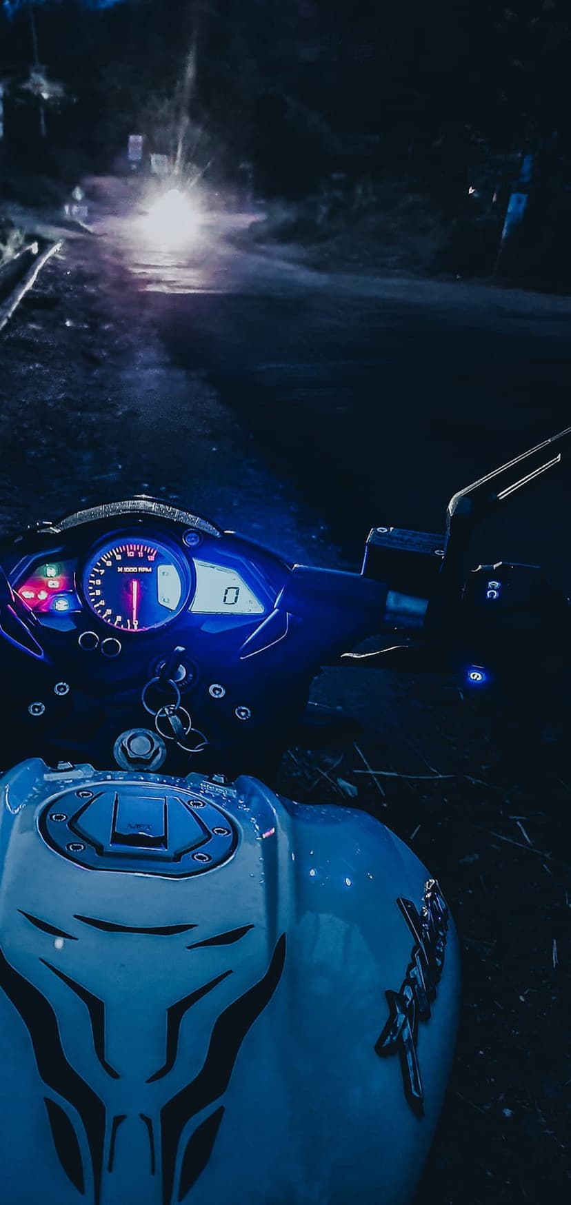 Motorcycle cockpit with glowing blue gauges and a tribal fuel tank view of a wet road at night from a rider POV.