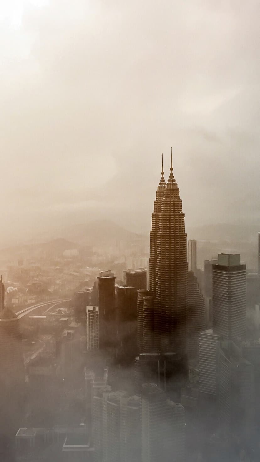 Twin towers with pointed spires pierce through thick urban mist in a warm sepia-toned cityscape view.