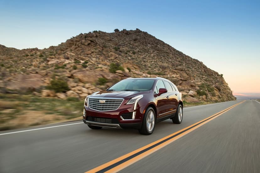 A dark red Cadillac SUV drives on an asphalt desert road past a large rocky hill under a clear sky.
