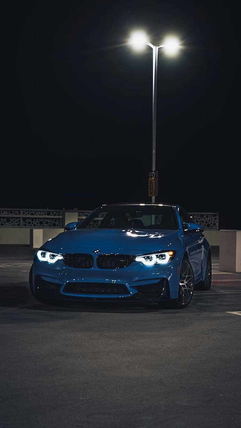 Blue BMW M4 parked under a bright streetlamp at night with illuminated headlights and a dark industrial backdrop.
