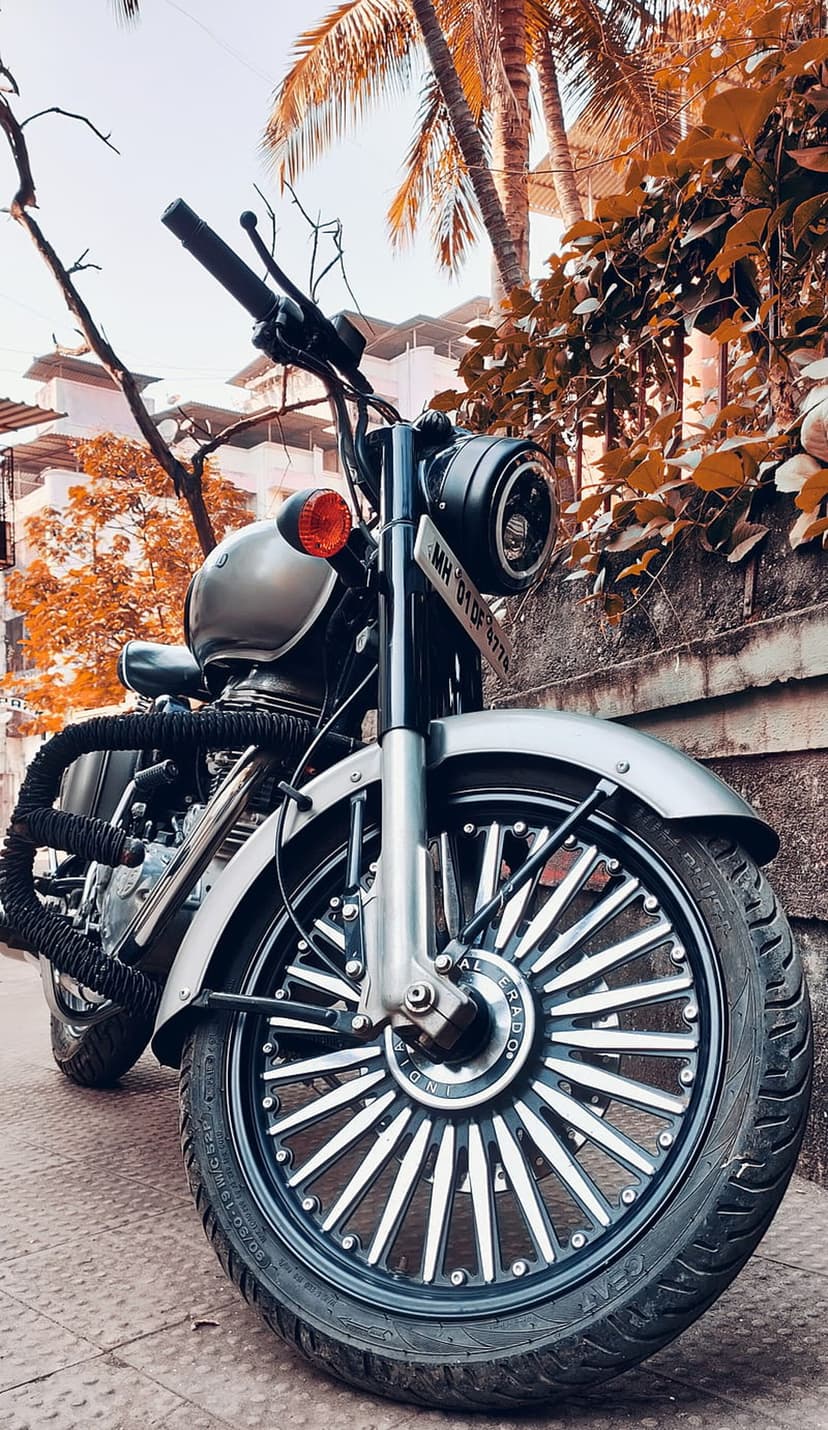 Classic silver motorcycle with custom spoke wheels parked near a stone wall and colorful autumn foliage.