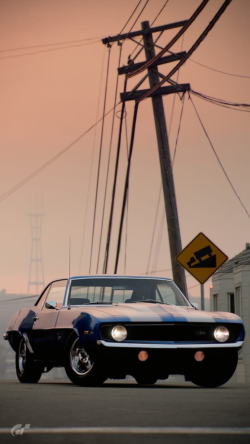 Blue vintage Chevrolet Camaro parked on a road with overhead power lines and a yellow steep grade sign at sunset.