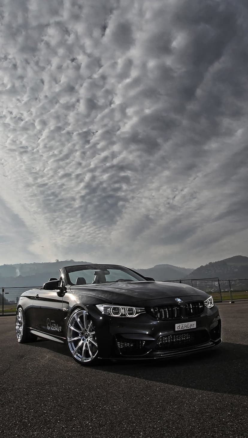 Black BMW M4 convertible seen from a low angle under a dark, cloudy sky with custom wheels and sleek bodywork.