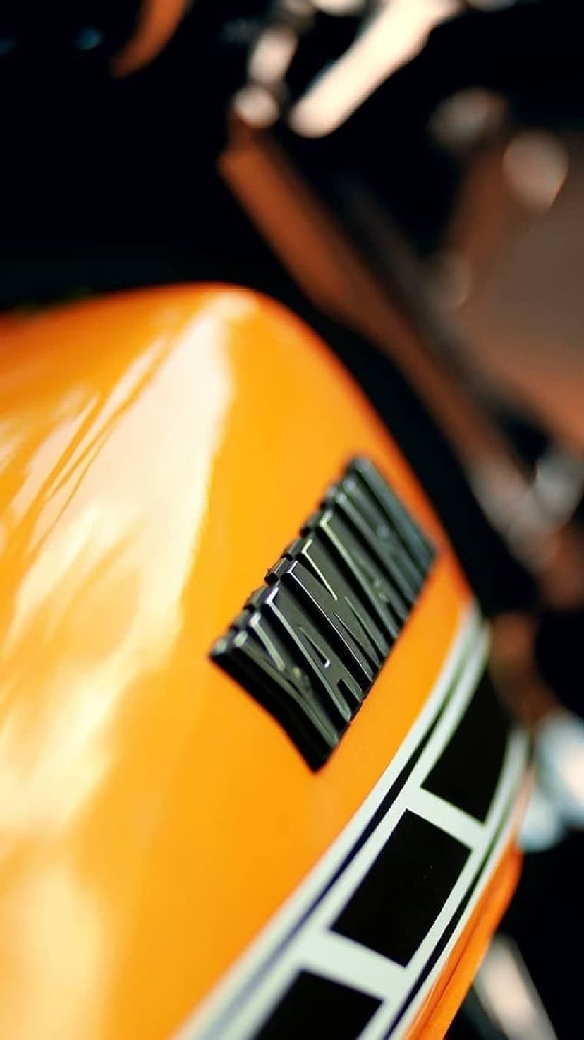 Yamaha chrome emblem and black racing stripes on a vibrant orange metallic motorcycle fuel tank close-up.