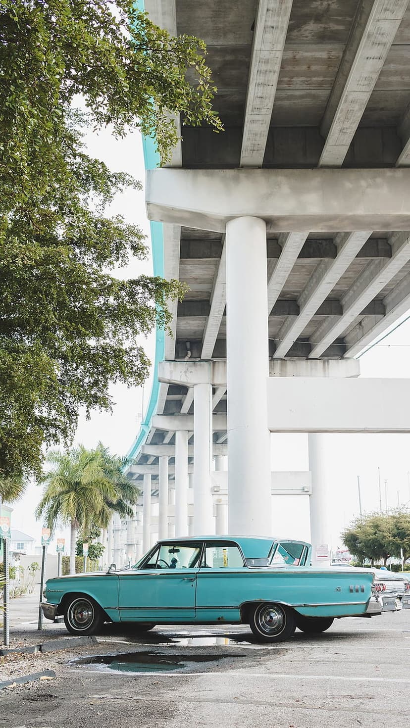 Vintage teal car parked on asphalt under a concrete bridge with palm trees and a cloudy sky in the background.