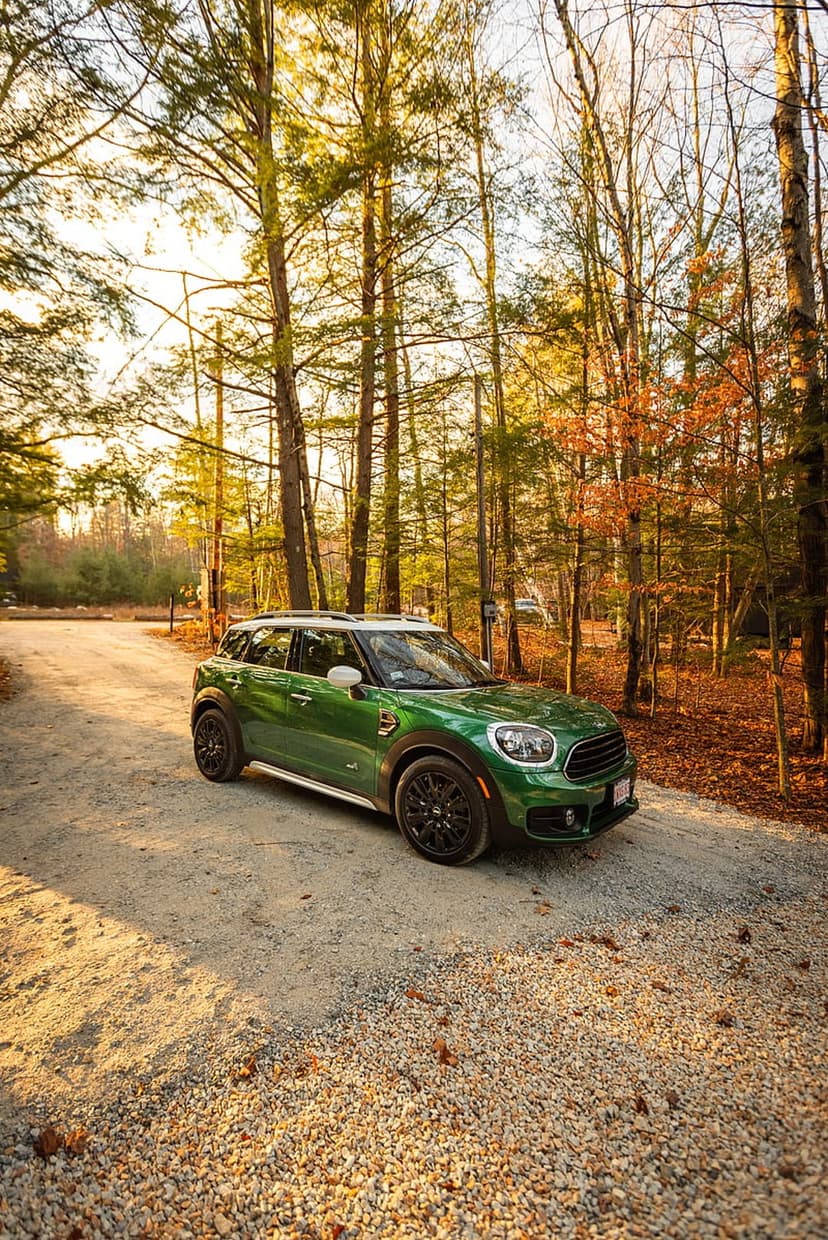 Green Mini Cooper Countryman parked on a gravel forest road with orange autumn leaves and golden hour sunlight.