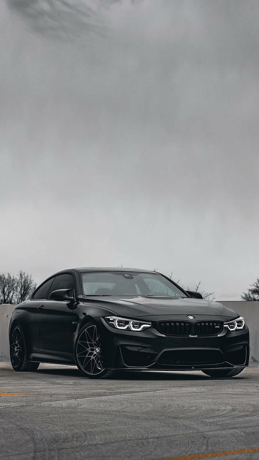 Matte black BMW M4 coupe with illuminated LED headlights parked on concrete against a cloudy, dramatic sky.