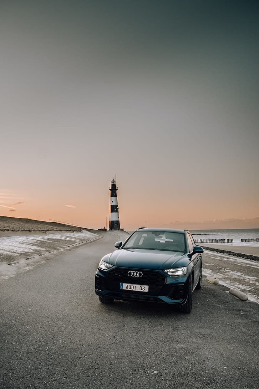 Audi Q3 SUV with headlights on parked next to a black and white striped lighthouse on a coastal road at dawn.