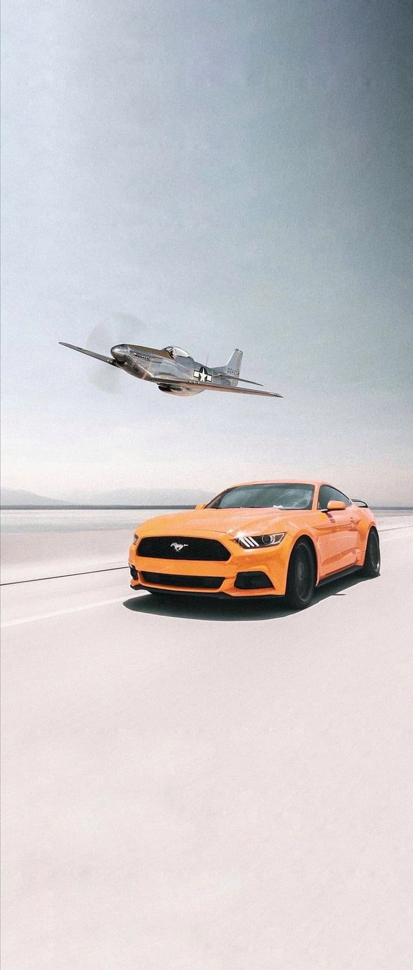 Orange Ford Mustang car driving on a road below a silver P-51 fighter plane flying in a bright blue sky.