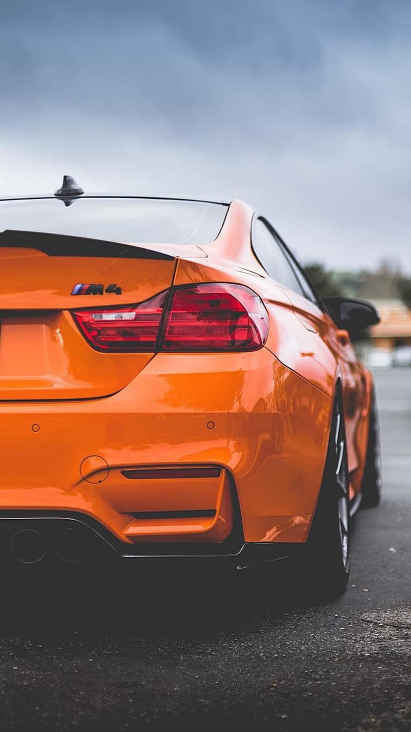 BMW M4 rear taillights and trunk badge on a bright orange sports car parked on dark asphalt under a gray sky.