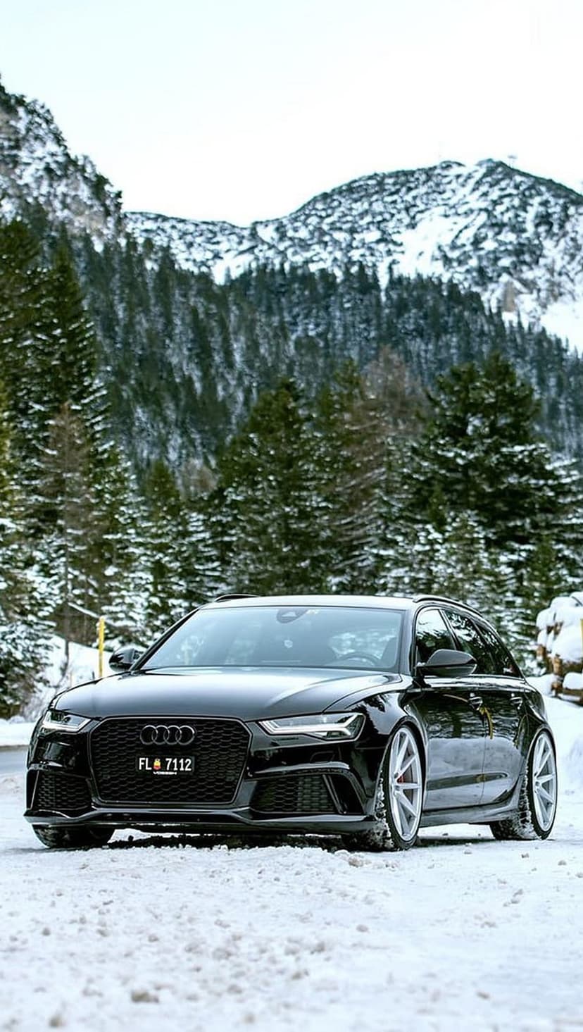 Black Audi RS6 Avant parked on a snow-covered mountain road surrounded by tall pine trees and winter peaks.