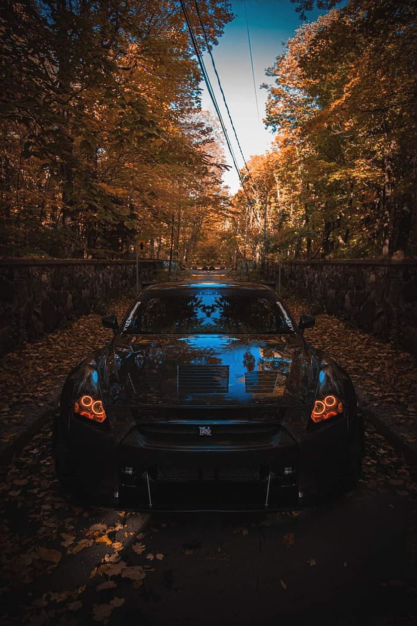 Nissan GT-R with glowing orange headlights parked on a rural forest road covered in fallen autumn leaves.