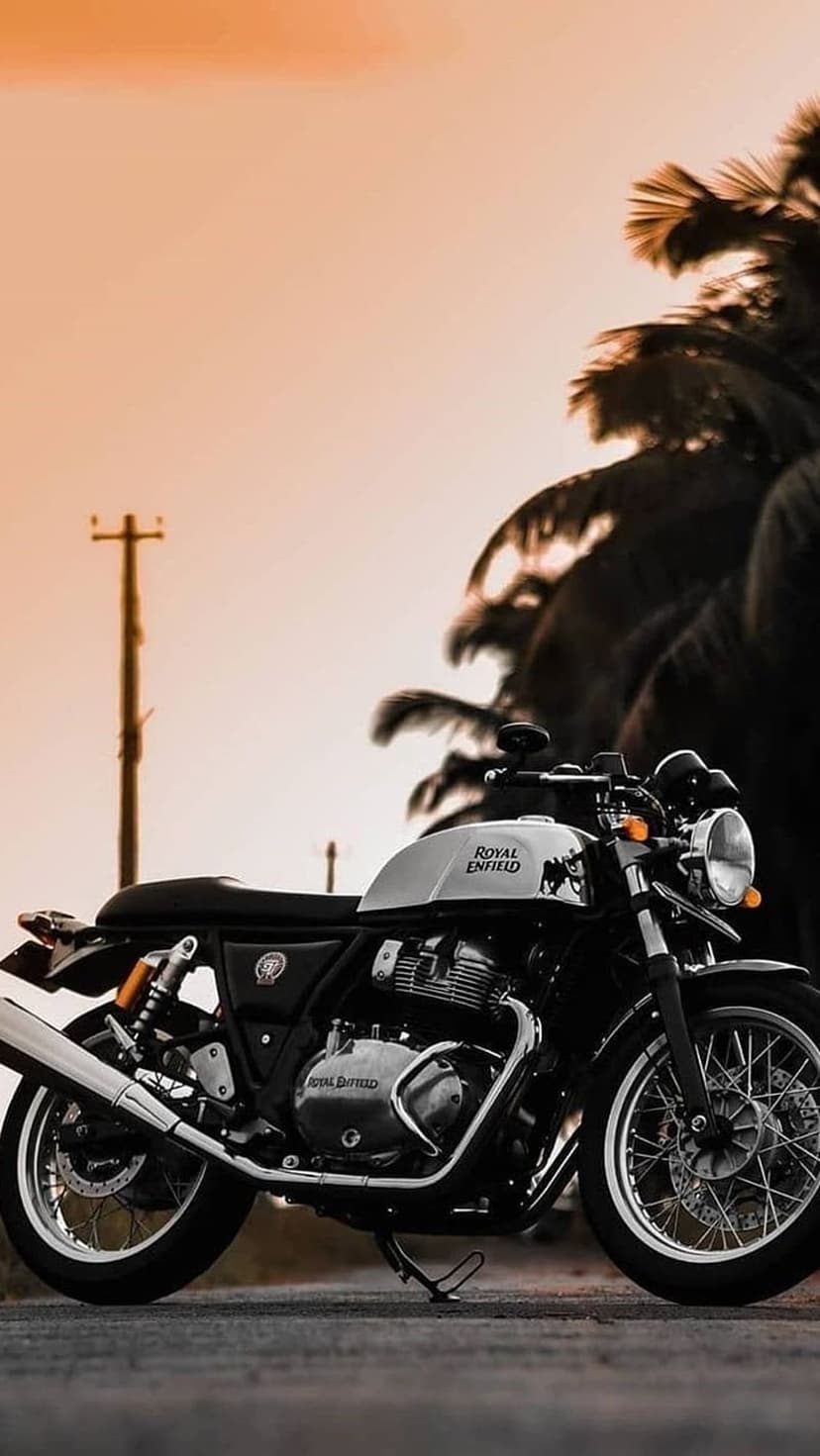 Royal Enfield motorcycle parked on a coastal road during an orange sunset with silhouetted palm trees.