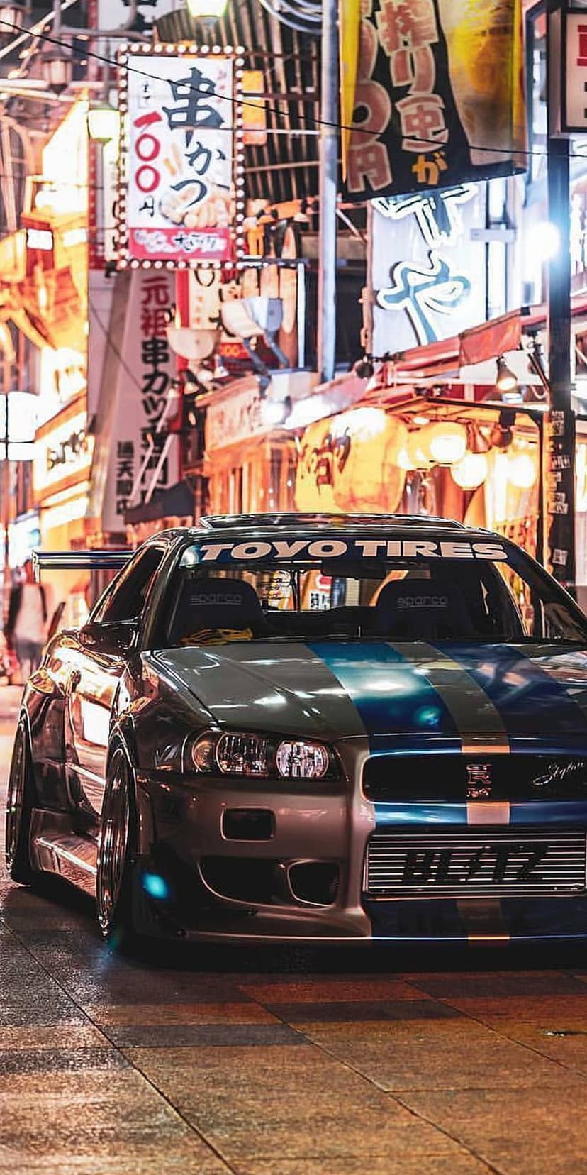Nissan GT-R R34 with blue livery parked on wet cobblestones under glowing neon Japanese city signs at night.