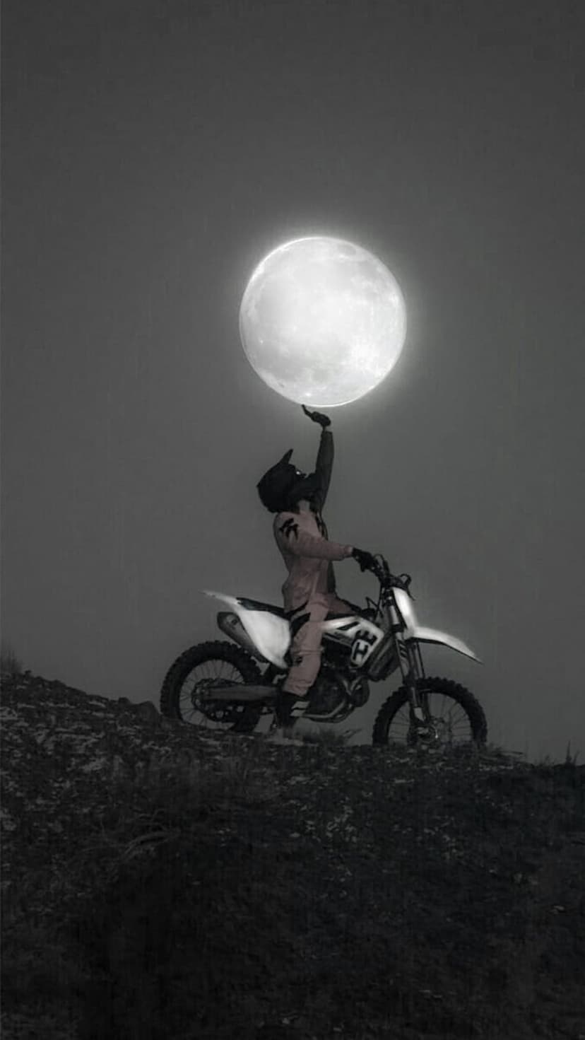 Motocross rider on a dirt bike reaches a hand toward a massive full moon under a dark, cloudy night sky.