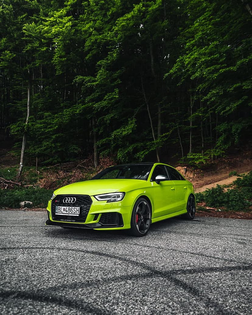 Lime green Audi RS3 sedan drifting on a paved road with tire marks against a dense green forest background.
