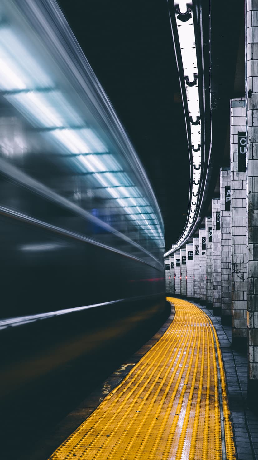 Metro train blurring past a curved subway platform with yellow tactile paving and bright overhead lights.