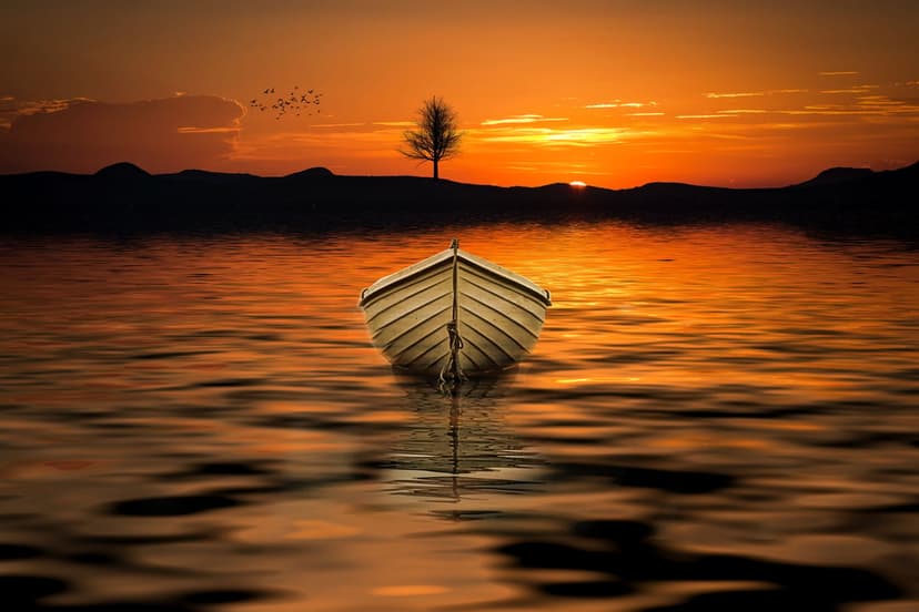 Small wooden rowboat on a still lake reflecting orange sunset clouds with distant hill silhouettes in the back.
