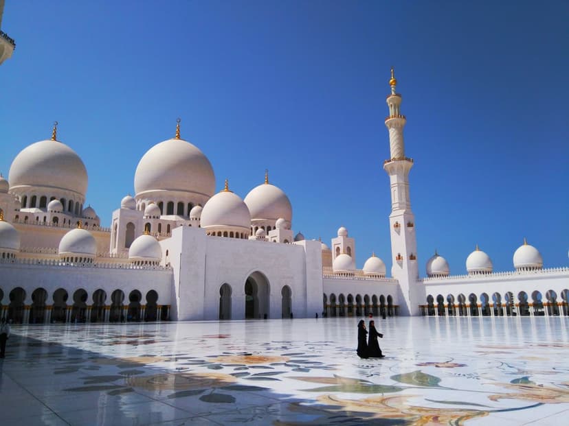 White marble mosque domes and a tall minaret reflect on a polished courtyard under a clear blue midday sky.