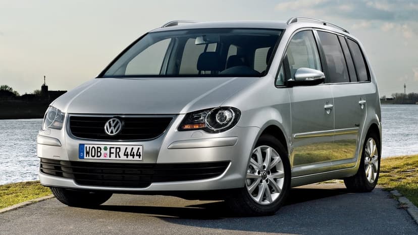 Silver Volkswagen Touran minivan parked on a paved path by a calm river under a clear sky in bright daylight.