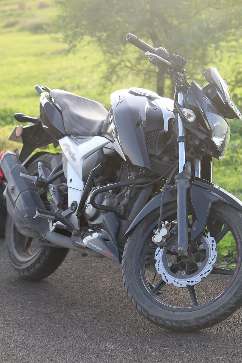 TVS Apache motorcycle with disc brakes parked on asphalt near a blurred green field under soft sunlight.