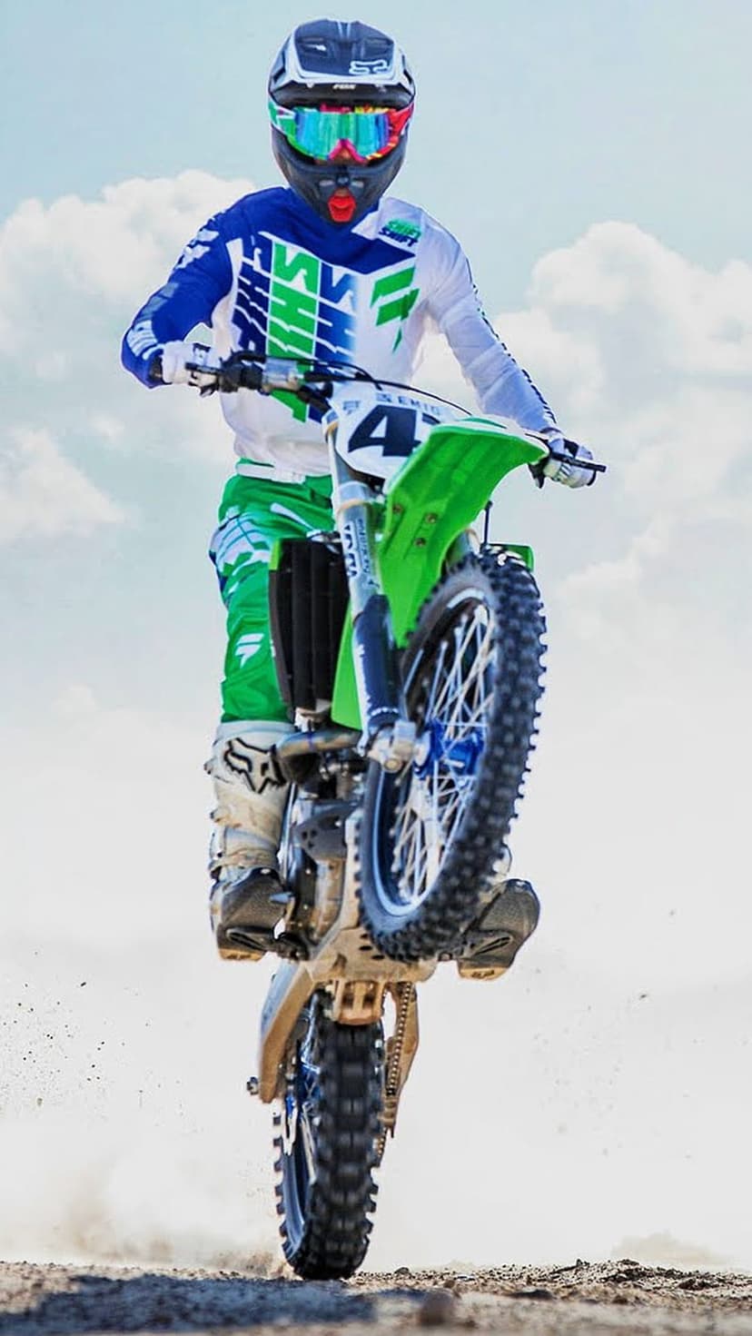 Motocross rider in a blue racing suit performs an aerial wheelie on a green dirt bike against a cloudy sky.