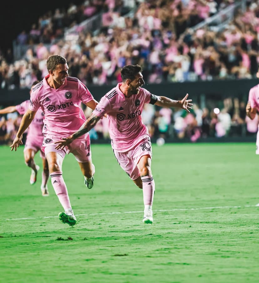 Inter Miami soccer players in pink kits celebrate a goal on a green grass field with a blurred crowd behind.