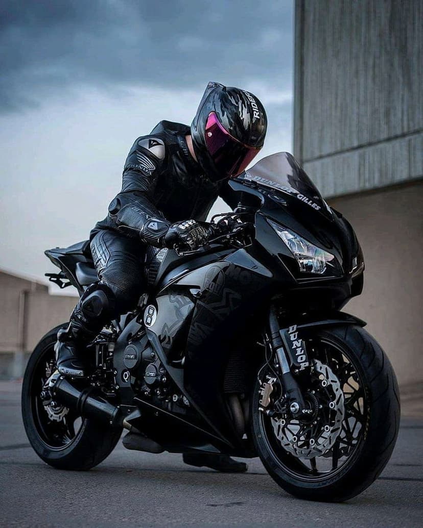 Motorcyclist in black gear with a pink visor sitting on a matte black sportbike against an urban backdrop.
