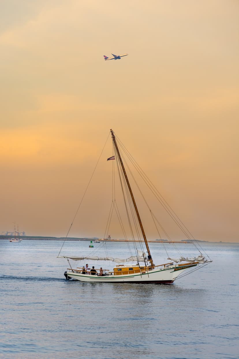Sailboat drifting on calm water under a distant airplane during a vibrant orange and yellow sunset horizon.