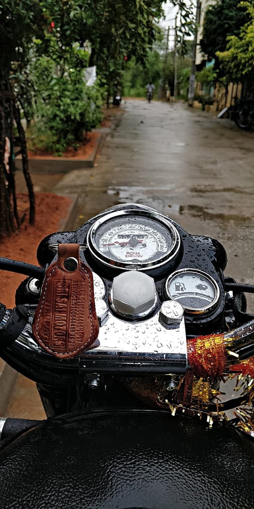 Motorcycle dashboard featuring a wet speedometer and fuel gauge before a blurred rainy road and green foliage.