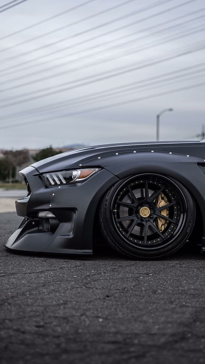 Matte black Ford Mustang with widebody fender flares, gold brake calipers, and a lowered stance on asphalt.