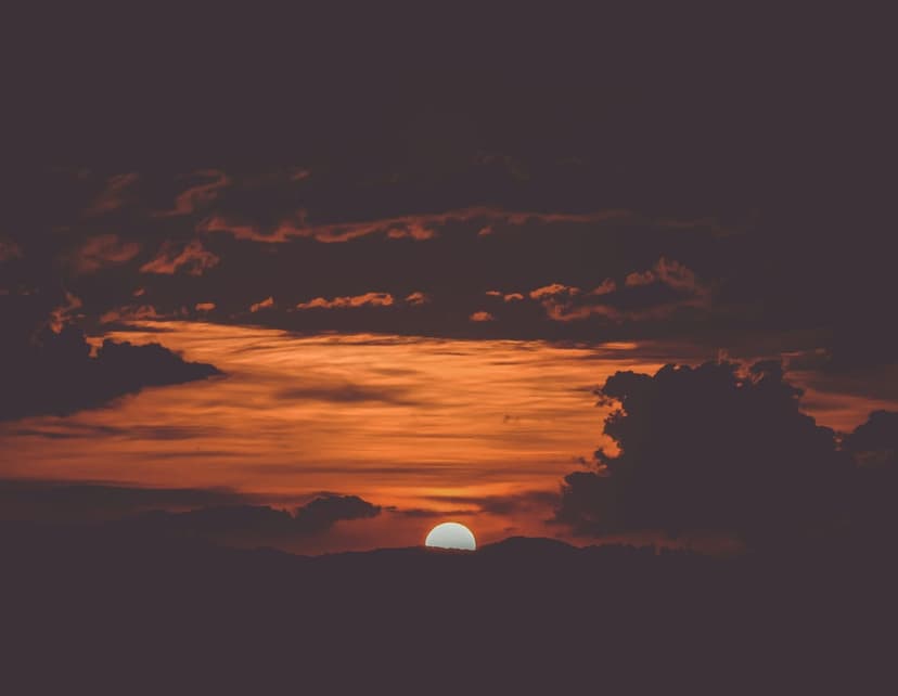 Glowing orange sun descends behind dark gray clouds during a vibrant twilight sky with deep red highlights.