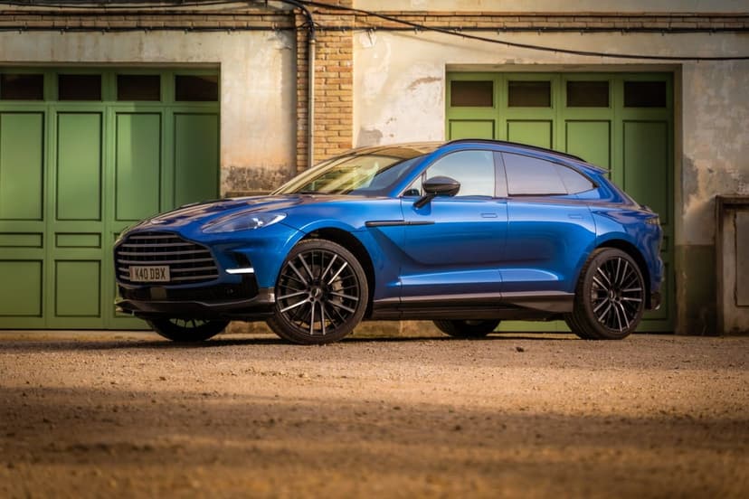 Blue Aston Martin DBX SUV parked on gravel in front of a weathered building with green double doors.