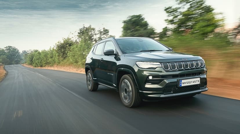 Dark green Jeep Compass SUV driving on a paved road with motion blur and forest background under a blue sky.
