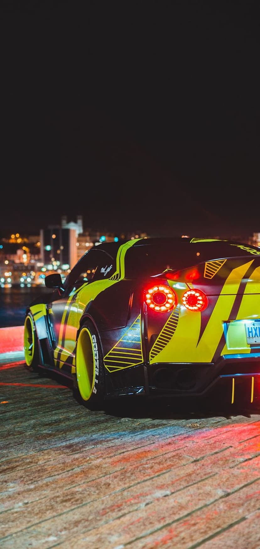 Yellow and black widebody Nissan GT-R parked on a wooden pier at night with a glowing city skyline background.