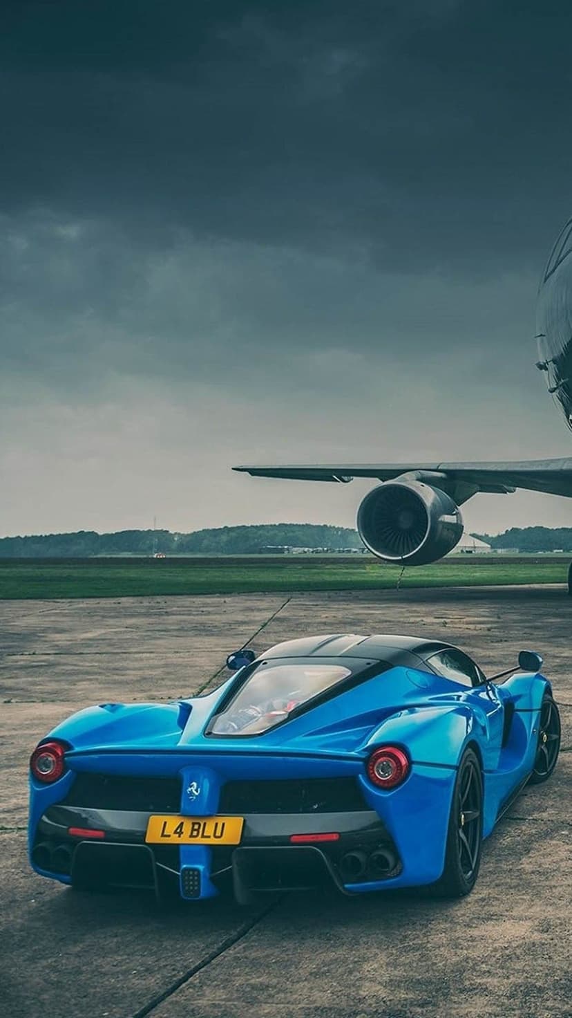 Rear view of a blue Ferrari LaFerrari on a runway beside a large jet engine under a dark, cloudy sky.