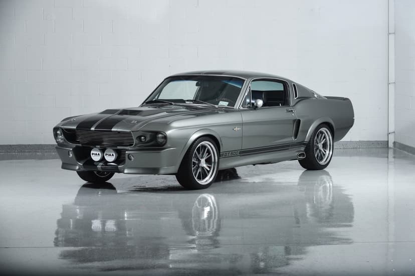 Silver Shelby GT500 Eleanor Mustang with black stripes parked on a reflective studio floor by a white brick wall.