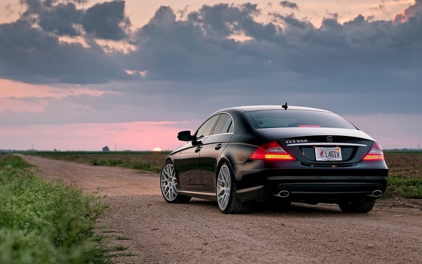 Black Mercedes CLS 550 on a dirt path under a pink sunset sky with glowing taillights and silver wheels.