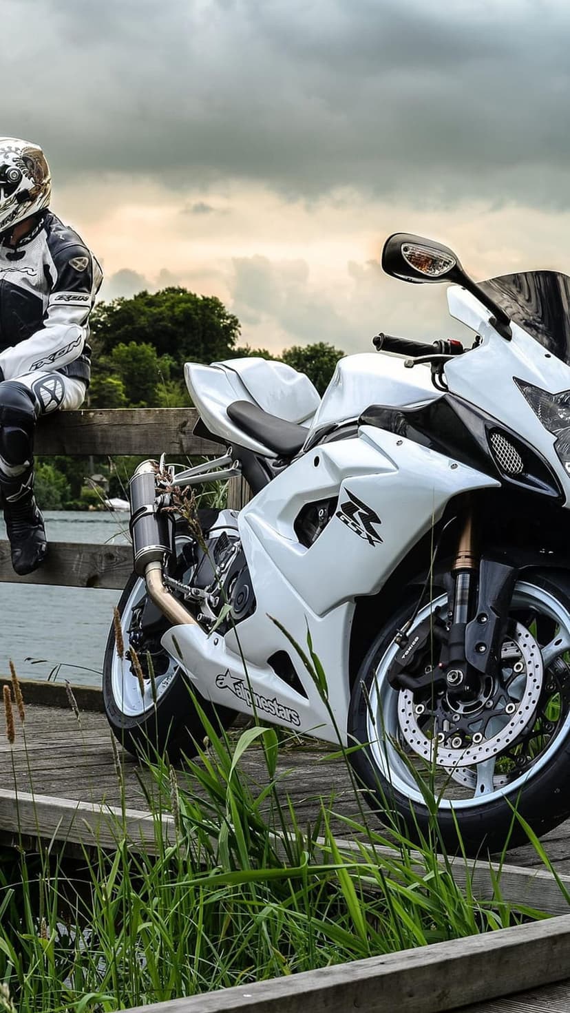 Motorcycle rider in full gear sits on a wooden dock next to a white sportbike overlooking calm lake waters.