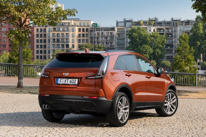 Rear view of an orange Cadillac XT4 SUV on cobblestones with city buildings and a blue sky background.