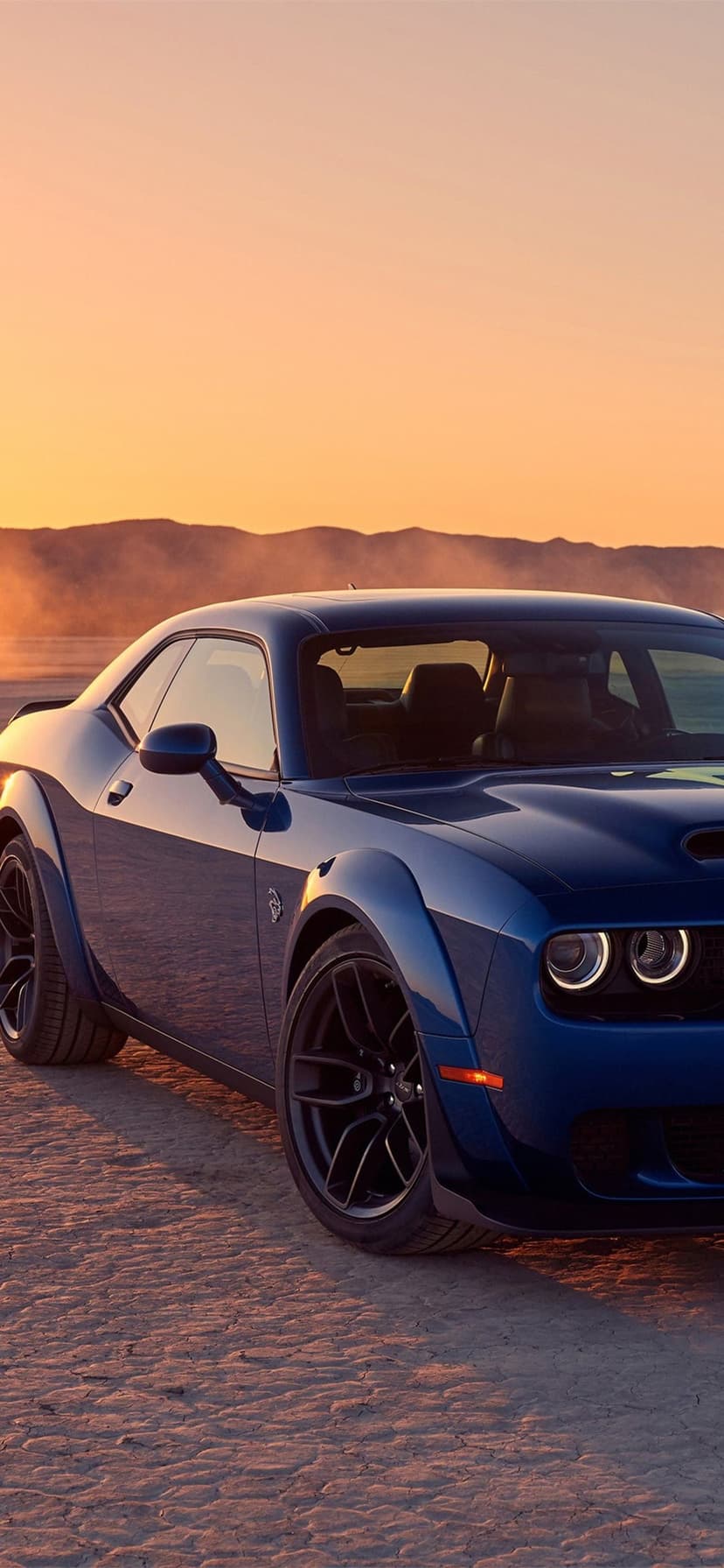 Blue Dodge Challenger Widebody on a dry desert lake bed under a glowing orange sunset sky with distant mountains.