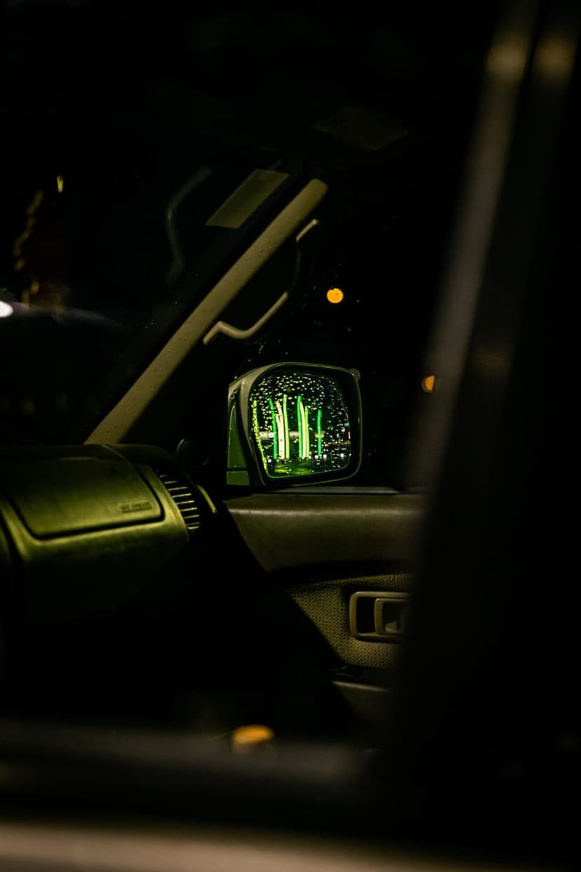 Car side mirror reflects glowing green digital patterns against a dark vehicle interior with distant orange lights.