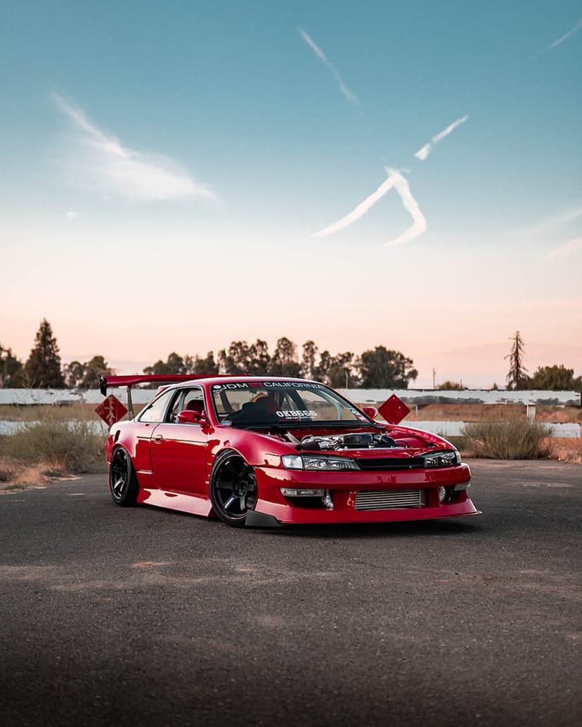 Red Nissan Silvia S14 drift car with an open hood and large rear wing stands on a dusty desert track at sunset.