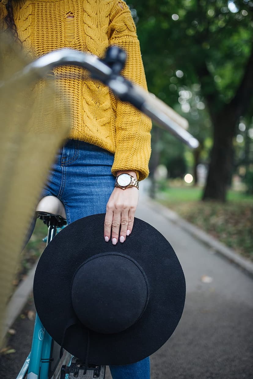 Woman in a yellow sweater holds a black hat beside a bicycle on a paved park path surrounded by green trees.